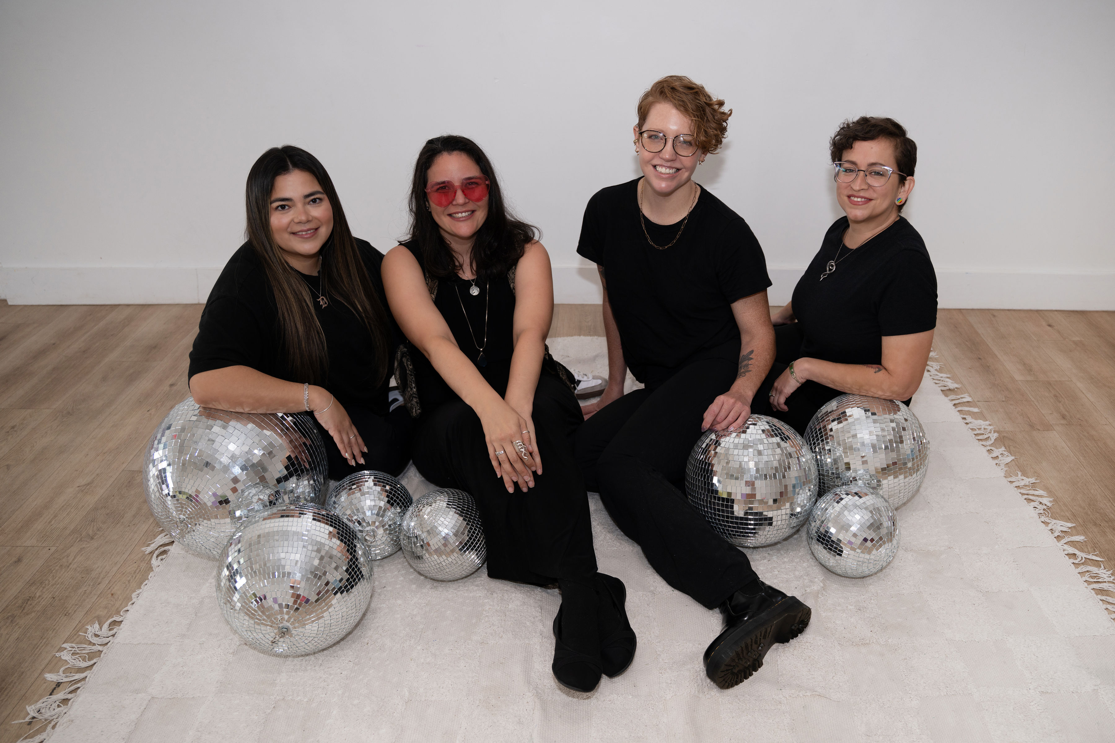 The Design Bodega team: four women in all-black seated together on the floor with silver disco balls in a bright white studio.