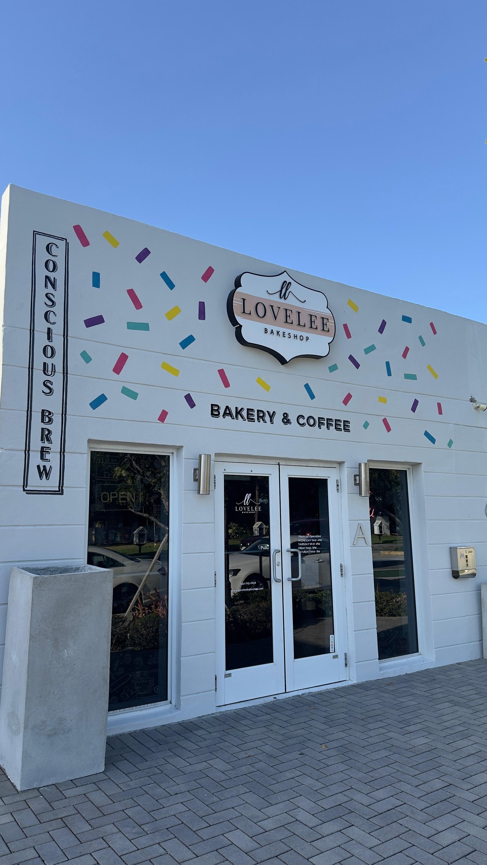 The storefront of Lovelee Bakeshop, a local business featuring a clean white exterior with colorful sprinkle decals and clear glass doors, representing the type of community-focused businesses Design Bodega partners with.